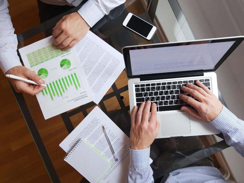 Overhead shot of two men, one using a laptop, and the other reviewing documents, business owners planning for tax interest deductibility changes