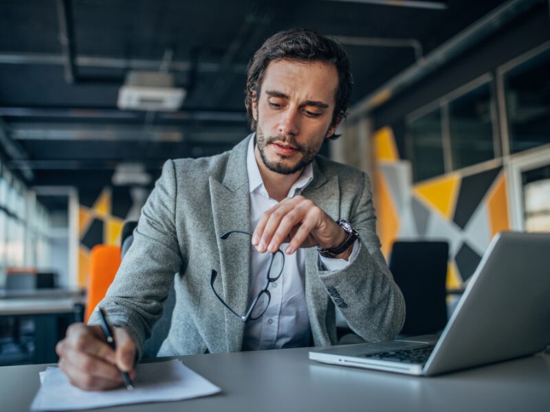 A man holding his glasses writes on a document with a slightly stressed expression, a business owner dealing with the policy changes surrounding the deductibility of ATO interest charges