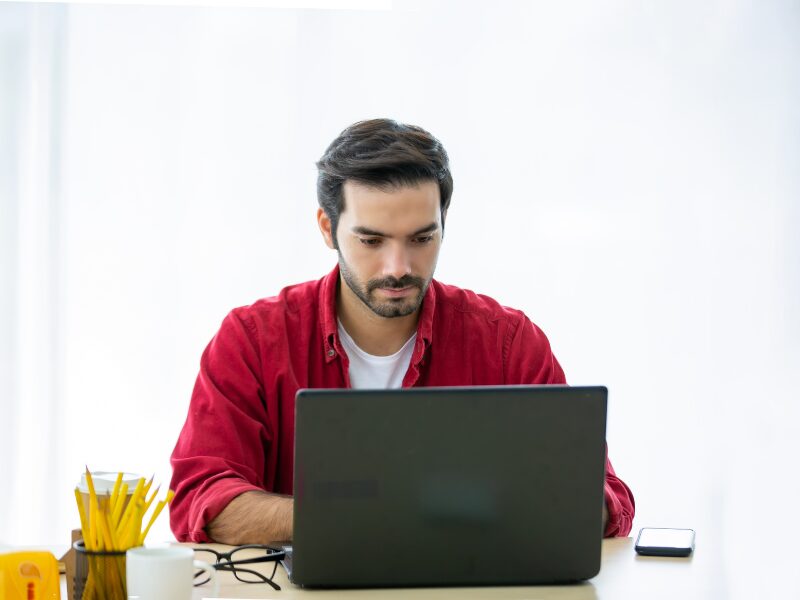 A man in a red shirt uses his laptop, a business owner preparing for ATO interest policy changes
