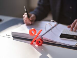A man writes on documents while using a calculator, a red 3D graphic of a percentage sign in front symbolising interest charges