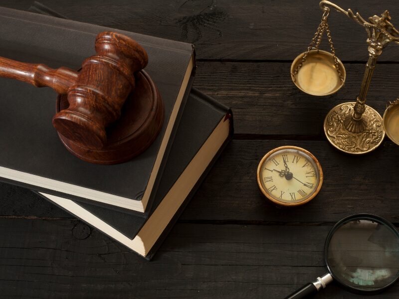 A judge’s gavel on top of two black books, an old-fashioned clock, a magnifying glass, and an ornate law scale placed on top of a dark wooden table