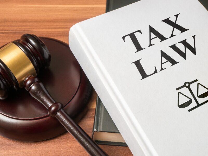 A large white book with the text “Tax Law” and a law scales symbol piled on top of a grey book next to a judge’s gavel