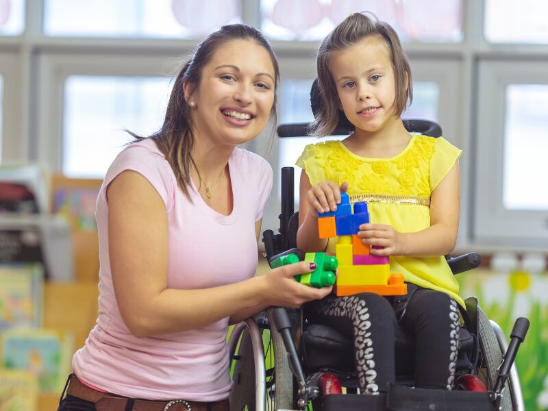 A disability support worker smiles with a child in a wheelchair while holding playing blocks