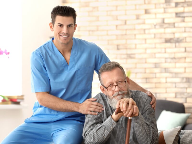 A disability support worker poses for a photo with his client, an elderly man in glasses holding a cane