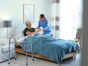 A disability support worker hands a glass of water to an elderly woman sitting in bed while reading a book, with a walker placed beside the bed