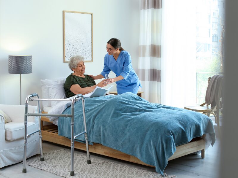 A disability support worker hands a glass of water to an elderly woman sitting in bed while reading a book, with a walker placed beside the bed