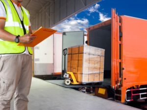 A freight company worker with a clipboard oversees a shipment being loaded into an orange freight truck