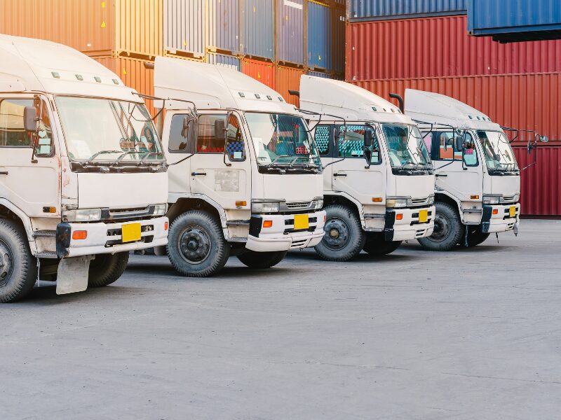 A row of four white freight trucks parked in front of shipping containers