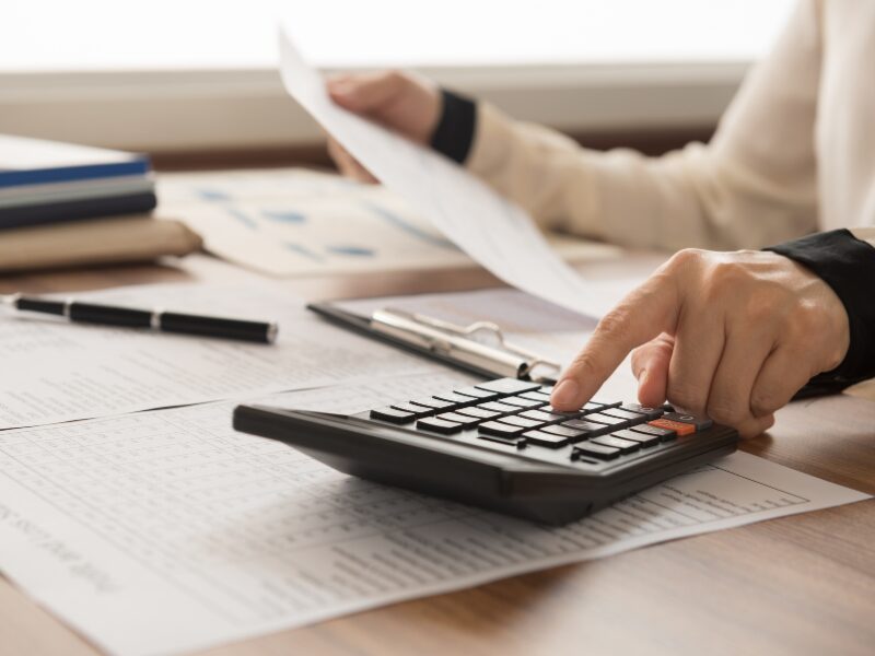 Cropped photo of a person showing only their hands, one hand holding a document and the other using a calculator, documents, books, and a pen visible on the table