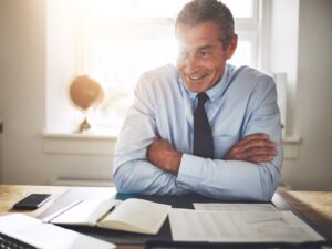 A financial planner smiles as his arms are folded, a notebook, pen, phone, and documents placed on the table, with bright light from the window behind him