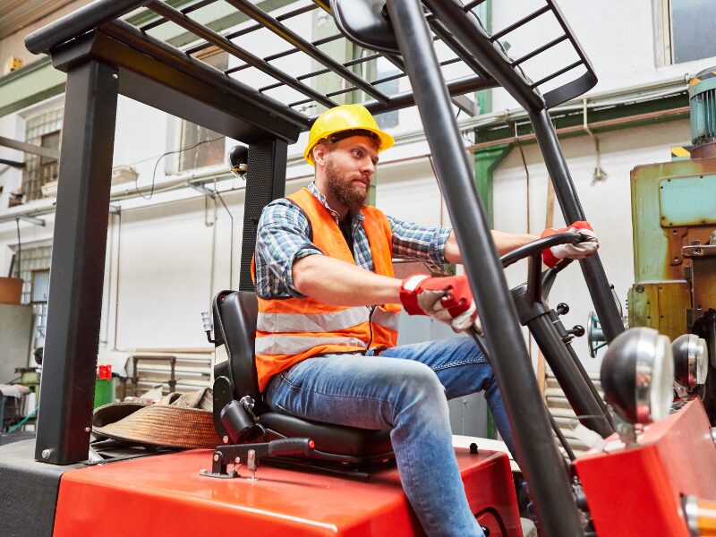 A construction operator wearing a hard hat and a high-visibility vest drives a small forklift