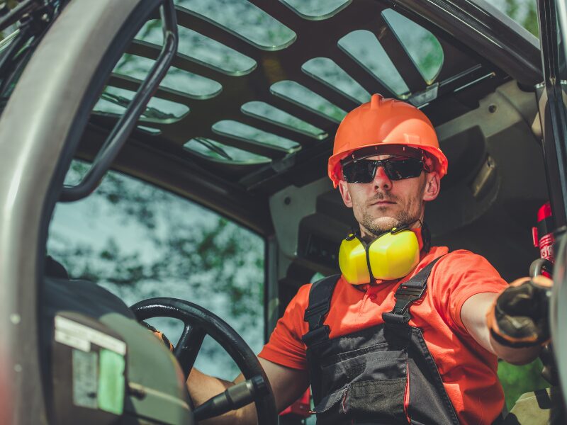 An equipment operator wearing a hard hat and sunglasses drives his equipment with a serious expression