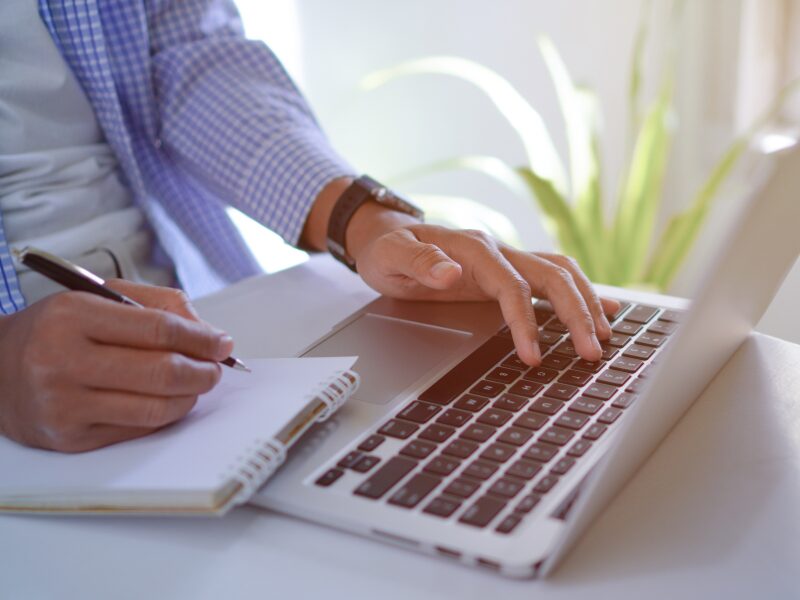 Cropped photo of a man using his laptop while writing on a notepad