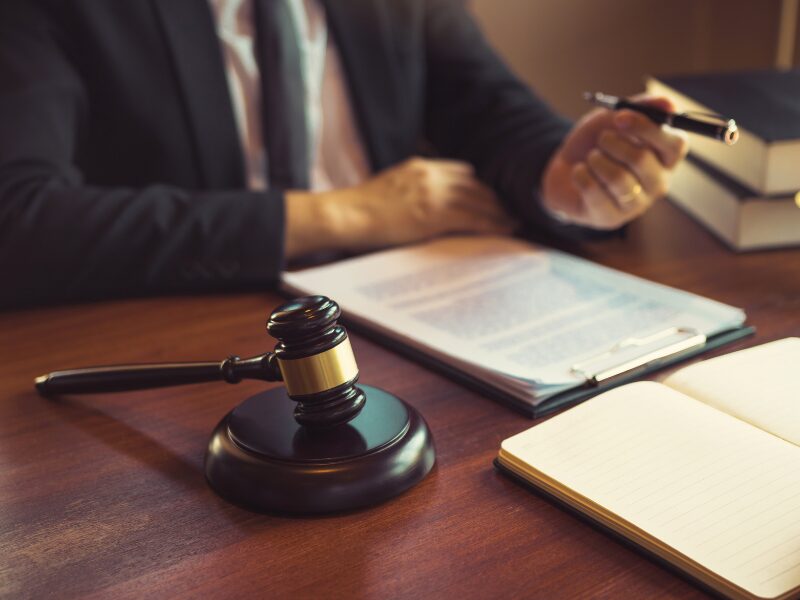 A man reads documents while holding a pen, a judge’s gavel in the foreground