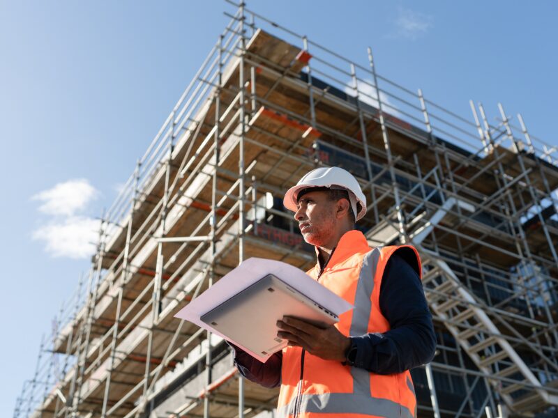 Low-angle photo of a civil construction worker holding a document and a laptop, ongoing construction project in the background
