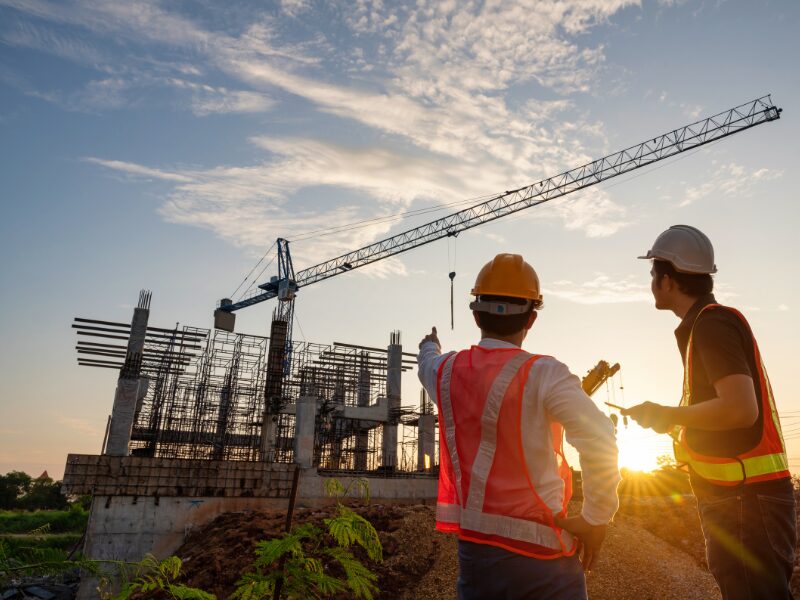Two civil construction workers in protective gear point at and discuss an ongoing construction project, the light from the sunrise shining between them