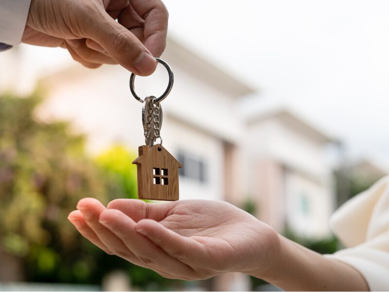 A hand giving a house key with a house-shaped keychain to another hand, new homes in the background