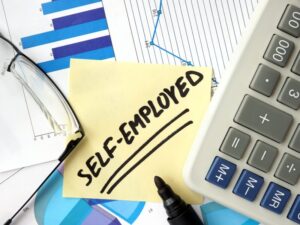 A zoomed-in photo of a yellow sticky note with the word “self-employed” underlined twice written on it, documents, glasses, a felt-tip pen, and a calculator surrounding it