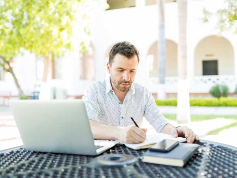 Self-employed man filling up a loan application at a table outdoors, laptop, notebook, and phone visible on the table
