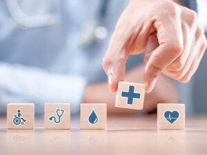 A hand places a wooden box with a medical cross symbol within a line of other wooden boxes with various medical-themed symbols on them, concept photo for allied health businesses