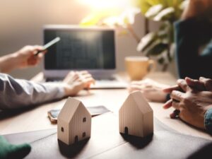 Two wooden miniature model houses in the foreground, the background shows a mortgage broker using a laptop to present refinancing loan details to clients