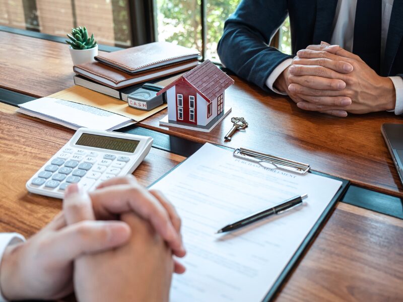 Cropped photo of a client and a mortgage broker or lender with both their hands clasped, a contract, pen, calculator, miniature model house, key, notebooks, and a stamp on the table between them