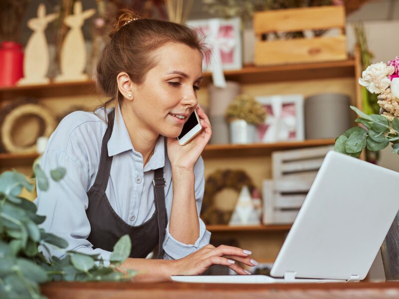 Small business owner wearing an apron, talking on the phone while working on her laptop