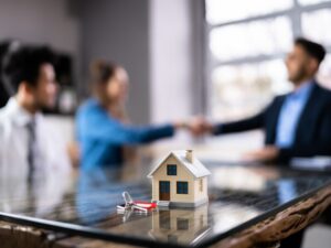 A miniature model house and house keys with a red keyring in the foreground, two home loan applicants shaking hands with a mortgage broker or lender in the background