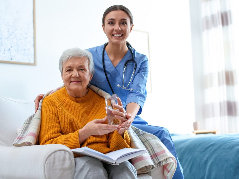 NDIS worker assists an elderly woman with holding a glass of water, looking at the camera and smiling