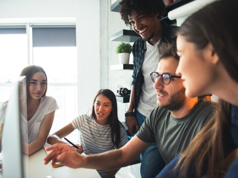 People working at a startup gathered at a computer while one of them discusses what’s on the monitor