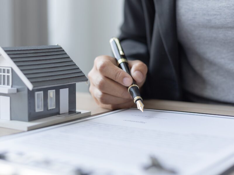 A man’s hand holds a fountain pen, ready to sign a self-employed home loan agreement. A miniature grey house is visible on the table