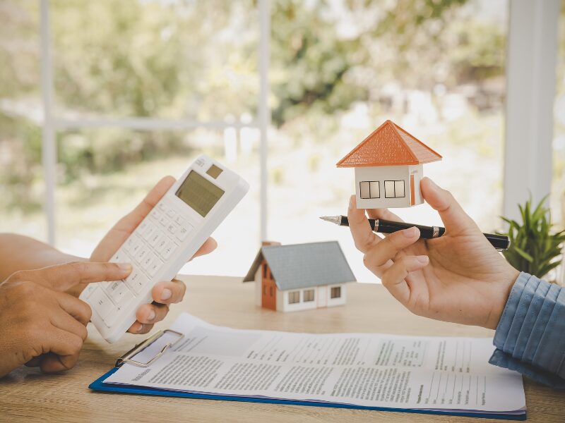 Cropped photo of two people in a discussion, showing only their hands, one person uses the calculator, one holds up a pen and a miniature model house, a document placed on the table, broker and borrower discussing using property as security for a loan