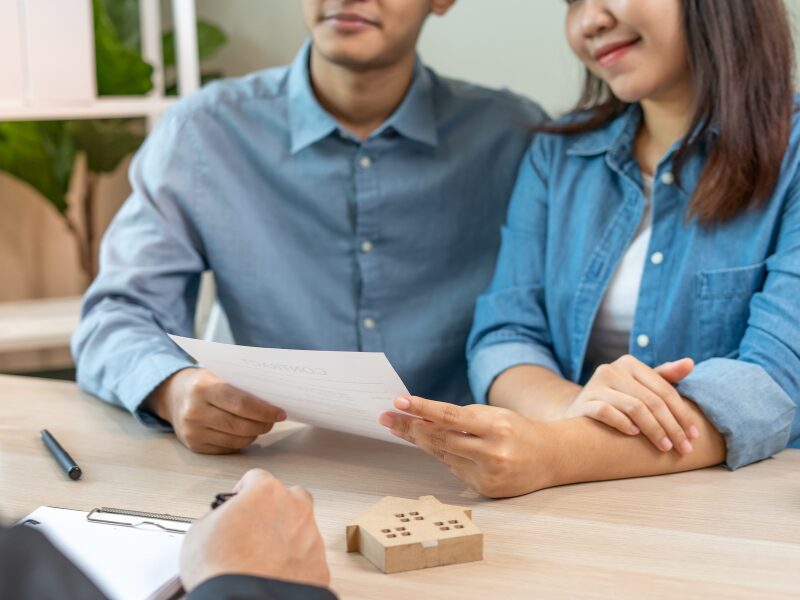 A couple reviews a caveat loan agreement while in a meeting with a loan broker or lender, a small wooden block shaped like a house is visible on the table