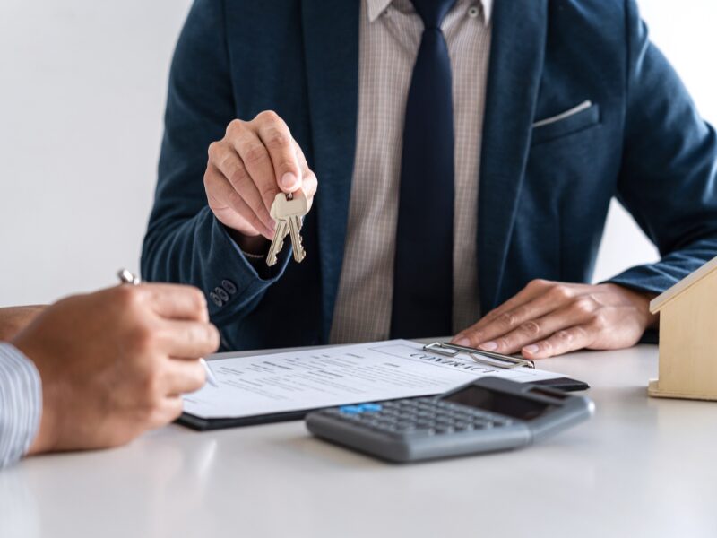 Cropped photo of a broker or lender handing over house keys to a self-employed applicant signing a loan agreement, a calculator and a miniature wooden house on the table