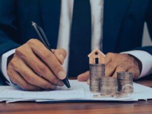 A man’s hand writes on a document, a pile of coins, a key, and a miniature model house beside the document, concept photo of a borrower leveraging property as equity for a caveat loan