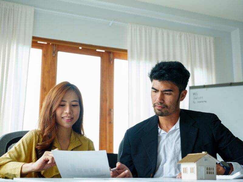 A broker discusses self-employed home loan eligibility criteria with a business owner, a woman discussing a document with a man, miniature model house visible on the table