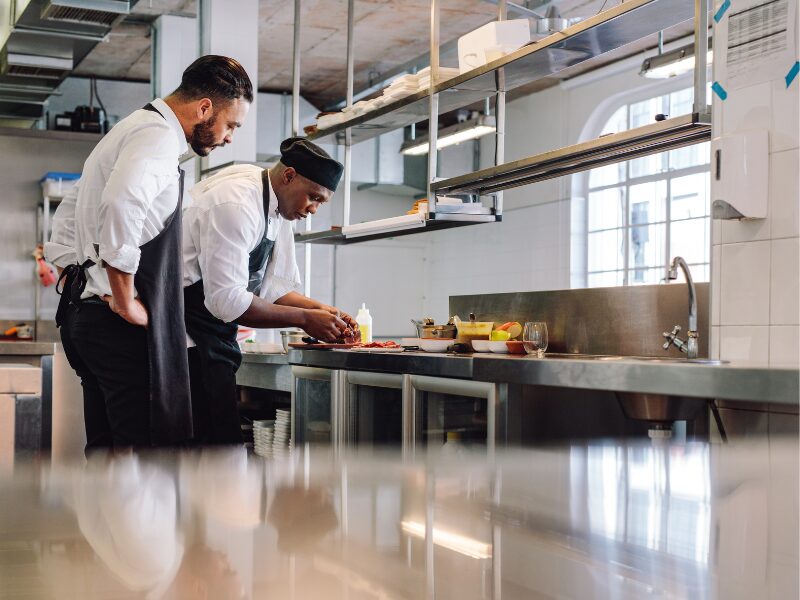 A chef prepares a meal at a commercial kitchen as another chef looks over and supervises
