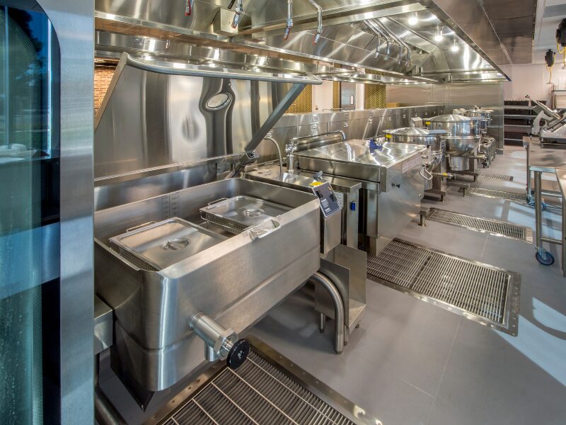 A clean, well-lit, and empty commercial kitchen with stainless steel sinks, drains, vats, and other equipment