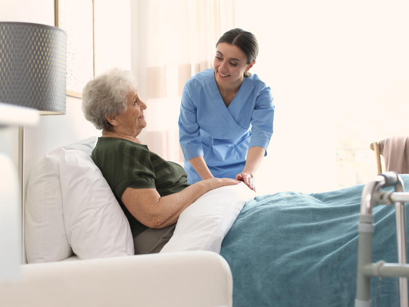 An NDIS worker happily checks up on an elderly patient sitting up on a bed