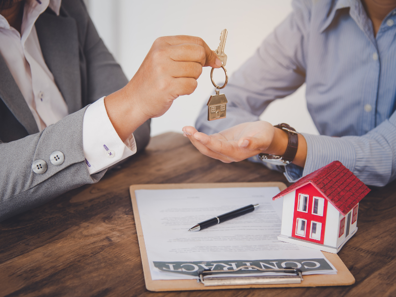 A mortgage broker or lender hands over the key to a new house to a self-employed borrower, a contract, pen, and a miniature house on the table