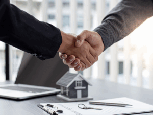 Two hands shaking, a loan contract, house key, pen, miniature house, and a laptop on the table below