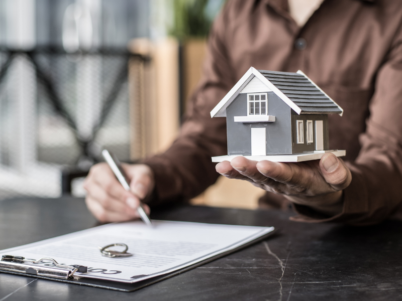 A man signs a loan contract while his other hand holds up a miniature model house