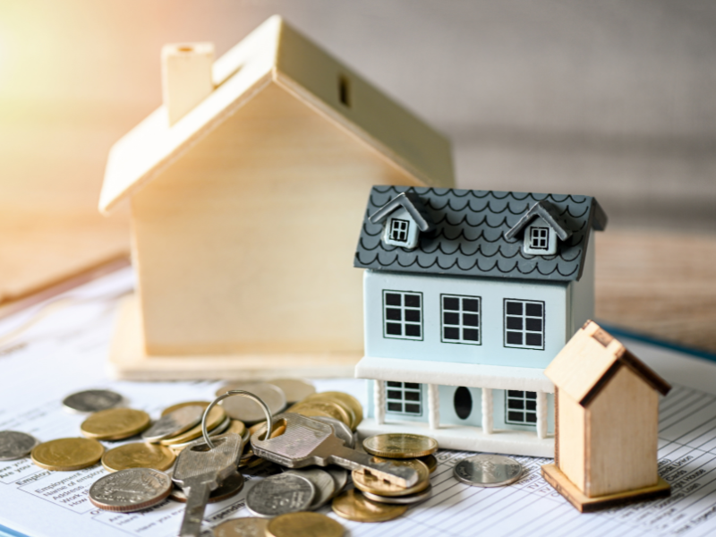 A pile of coins and house keys next to three miniature houses placed on top of a document