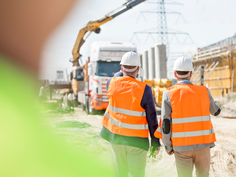 Two construction workers walking in a construction site, large equipment visible on the background