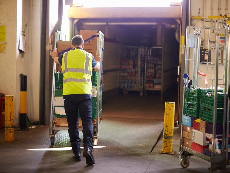 A worker in a high visibility vest pushes a cart full of boxed supplies into the car of a freight truck