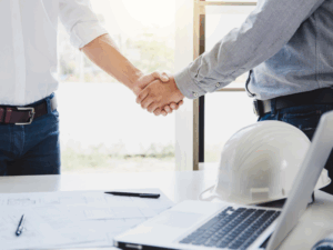 Cropped photo of two men shaking hands, building plans, a pen, a laptop, and a white hard hat placed on a table in front of them