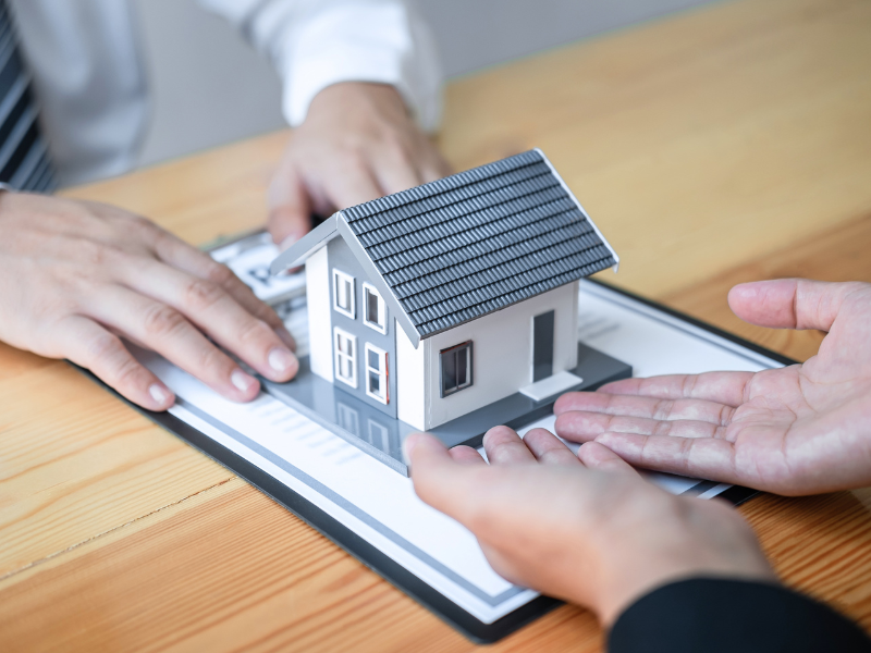 A pair of hands push a clipboard with a loan contract and a miniature house on top towards another pair of hands, concept photo for an approved low doc trust home loan