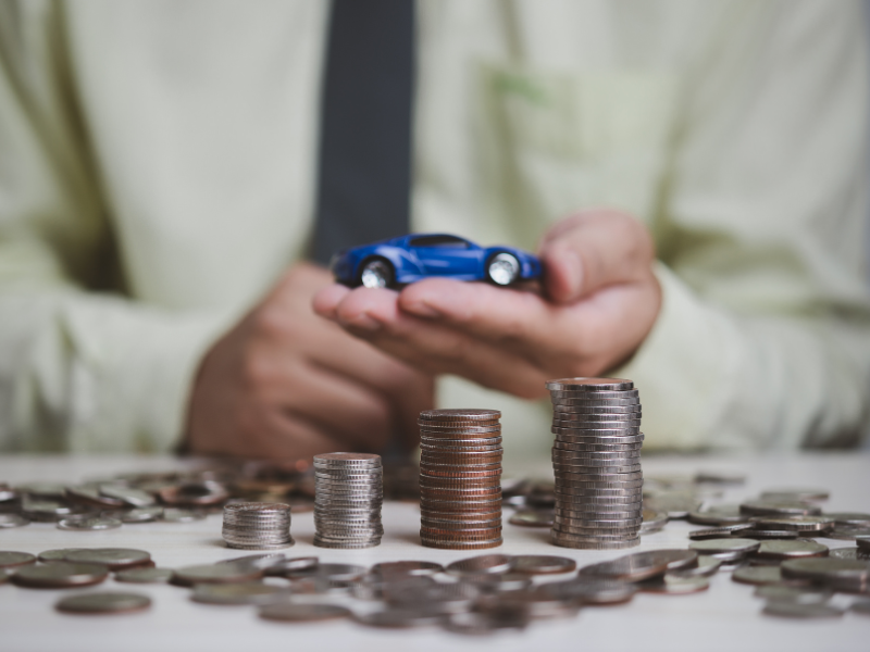 Cropped photo of a man holding a blue toy car, piles of coins on the table in front of him, concept photo for a car loan
