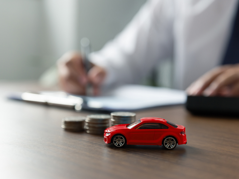 A red toy car next to a pile of coins, a man writing on a clipboard in the background, concept photo for a bad credit car loan