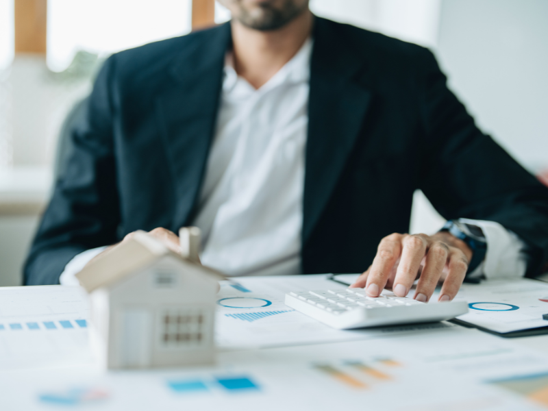 Cropped photo of a man in a suit using a calculator, a miniature house in the foreground, business owner calculating LVR for his low doc home loan